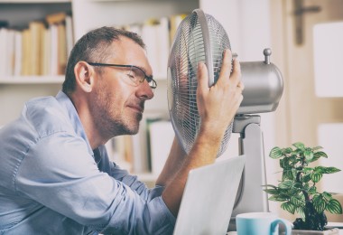 Mann sitzt im Büro vor einem Ventilator. Er hat die Augen zu und wirkt erschöpft.