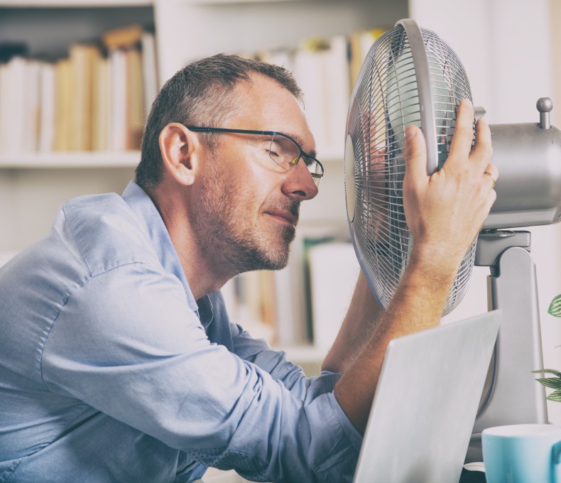 Mann sitzt im Büro vor einem Ventilator. Er hat die Augen zu und wirkt erschöpft.