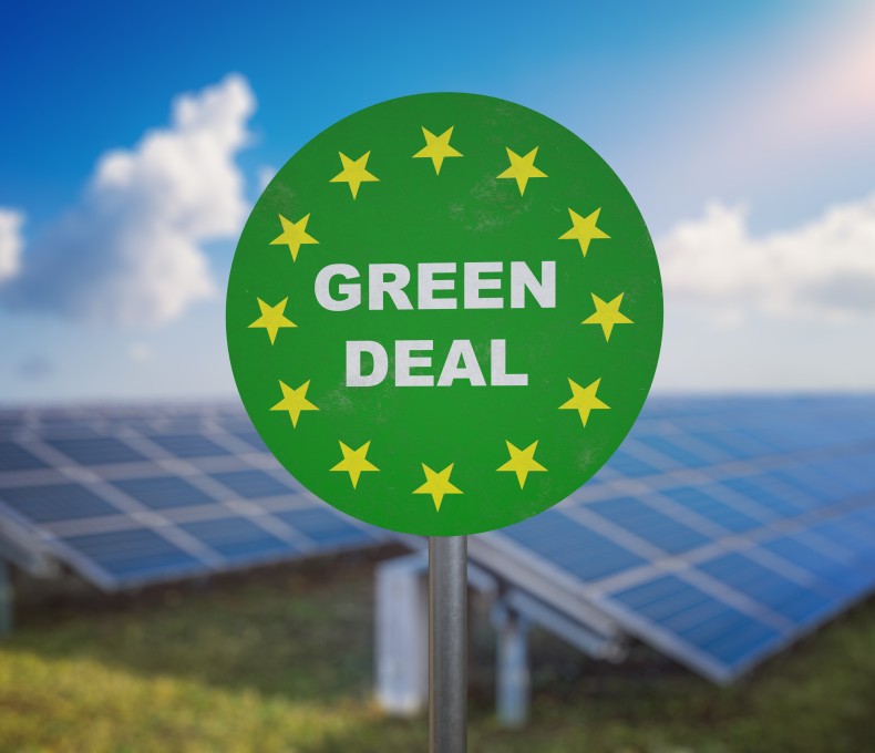 Auf einem grünen runden Schild steht: Green Deal; im Hintergrund sind Solarpanele zu sehen