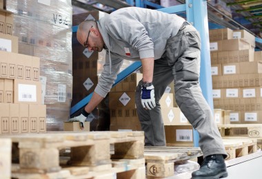 Person in Arbeitskleidung beugt sich in einem Lagerhaus über eine Holzpalette. Im Hintergrund sind Regale mit Kartons zu sehen.